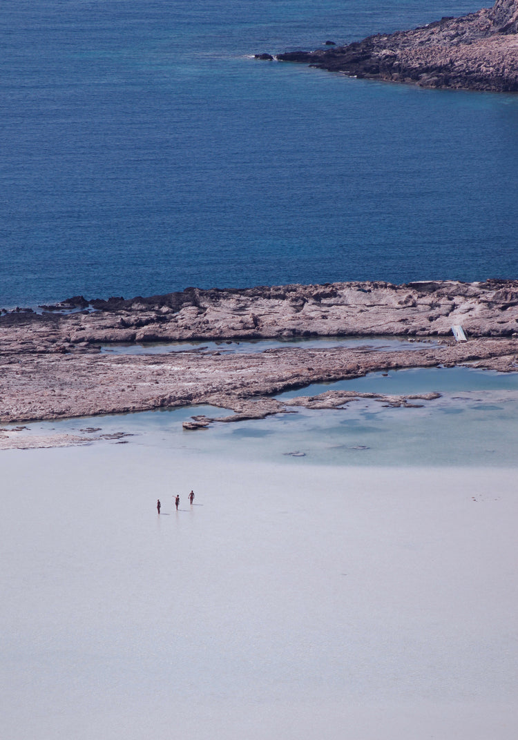 Three people walking in the shallows, heading towards the deep blue ocean ahead.