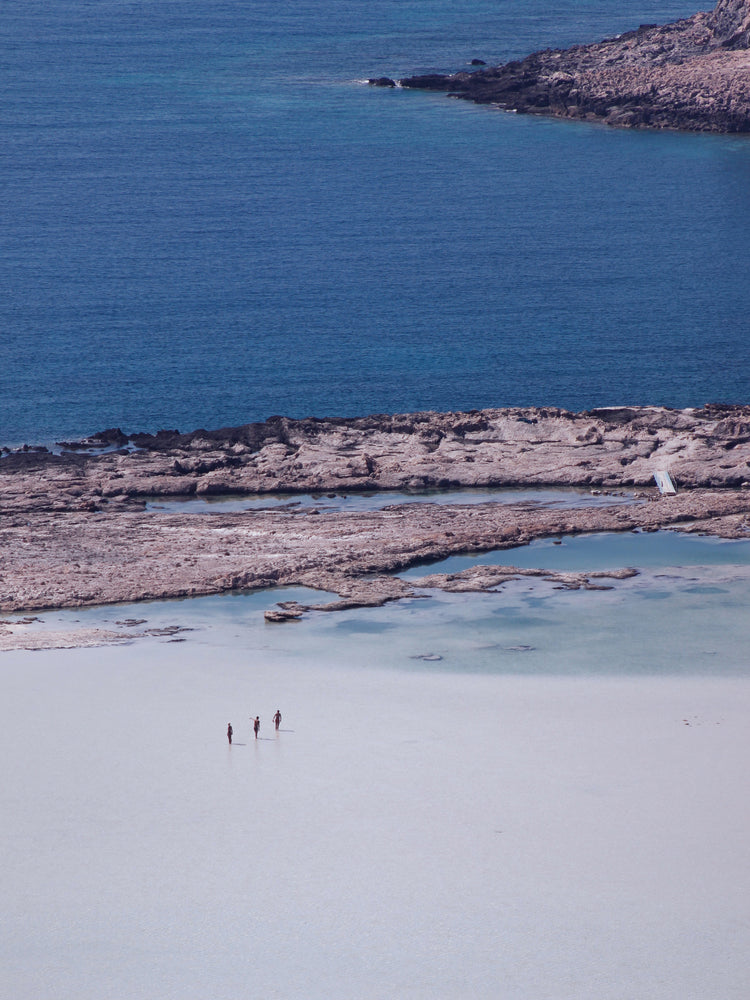Three people walking in the shallows, heading towards the deep blue ocean ahead.