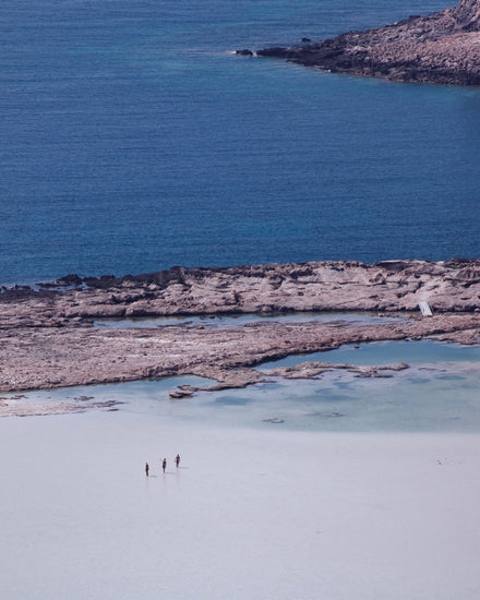 Three people walking in the shallows, heading towards the deep blue ocean ahead.