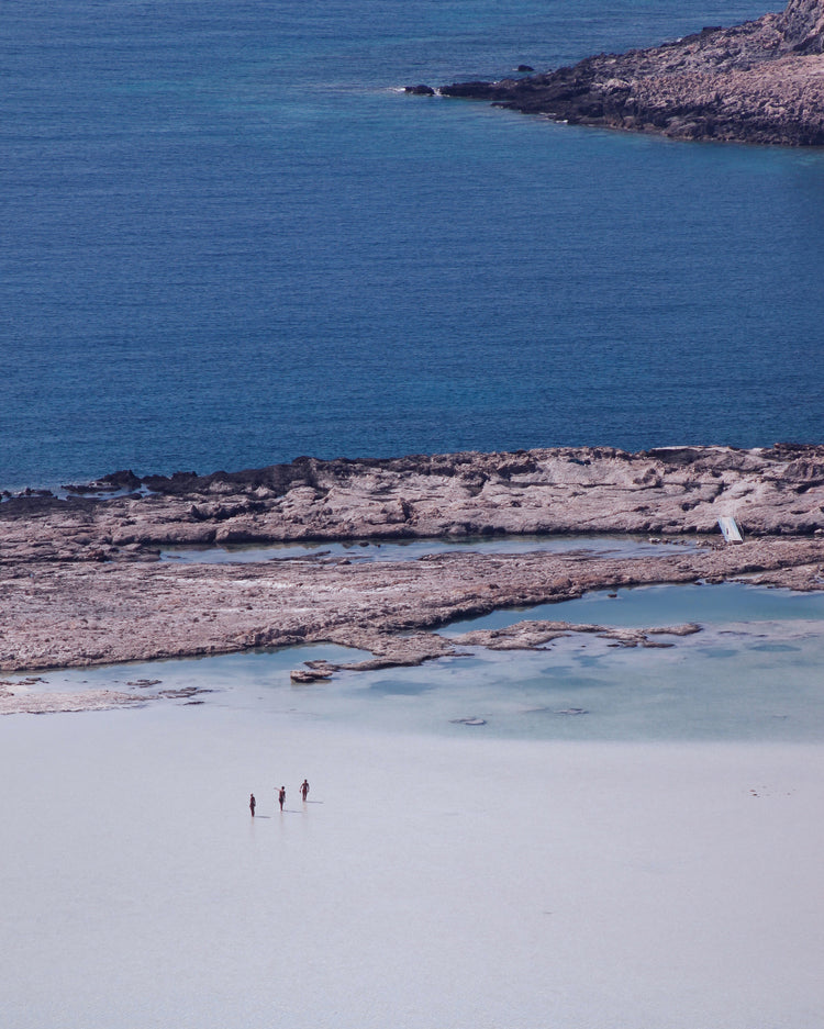 Three people walking in the shallows, heading towards the deep blue ocean ahead.