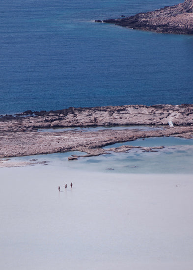 Three people walking in the shallows, heading towards the deep blue ocean ahead.