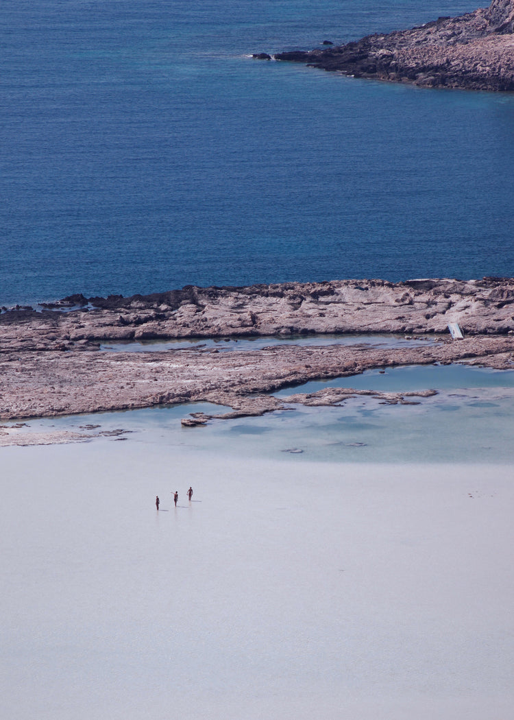Three people walking in the shallows, heading towards the deep blue ocean ahead.