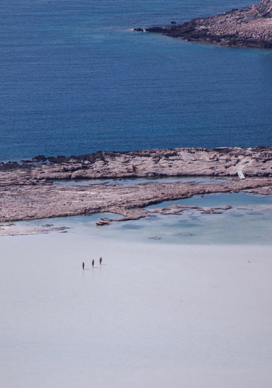 Three people walking in the shallows, heading towards the deep blue ocean ahead.