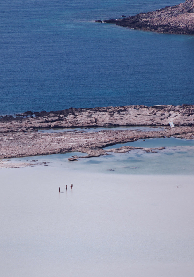 Three people walking in the shallows, heading towards the deep blue ocean ahead.