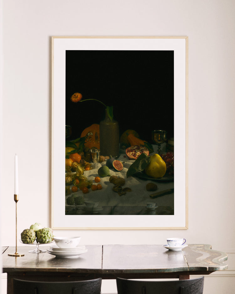 In a minimalist room, a wooden table adorned with two teacups, white plates, and artichokes is charmingly enhanced by a lit candle. Above, the Gloomy Picnic I Poster captures attention, blending classic art with subtle modernity reminiscent of Paul Cézannes style.