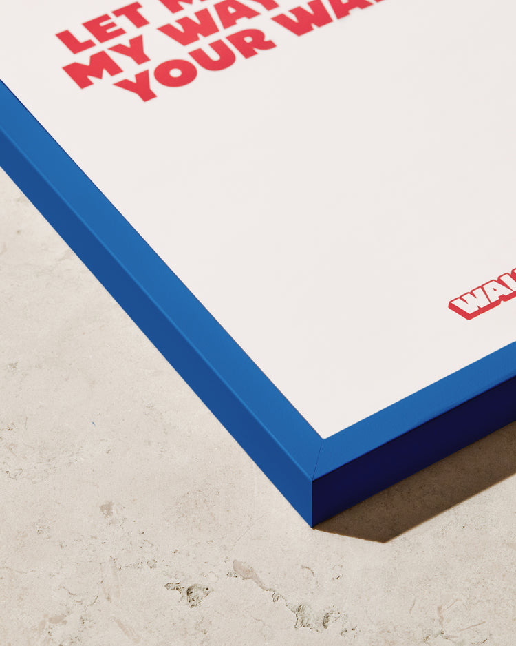 Close-up of the stylish Frame Blue poster in a blue wooden frame on a beige surface, featuring bold red text on a crisp white background.