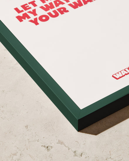 Close-up of a framed poster on a beige stone surface. The Frame Green adds a stylish pop with its dark green wooden frame, and the partially visible red text stands out against the white background.