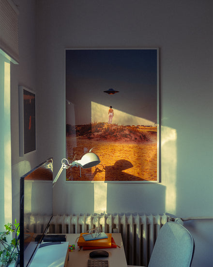 A cozy room with soft lighting has the Hey Luke, our Uber is Here! poster, depicting a person and UFO over a desert landscape, hung above a radiator. Nearby, the desk is adorned with a lamp, computer monitor, and office supplies.