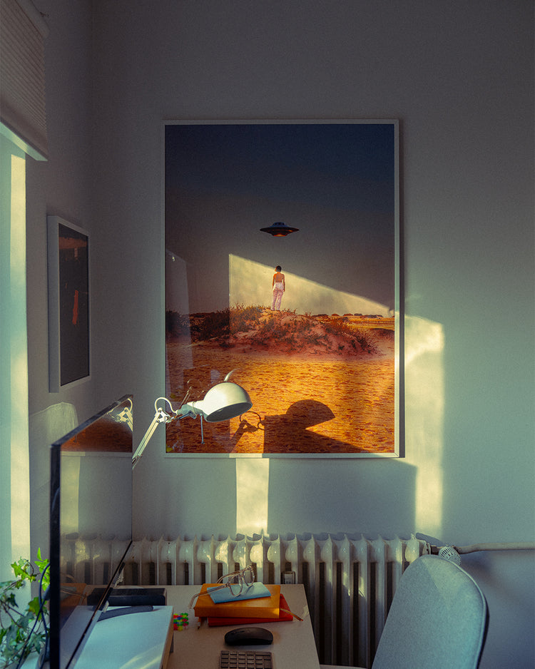 A cozy room with soft lighting has the Hey Luke, our Uber is Here! poster, depicting a person and UFO over a desert landscape, hung above a radiator. Nearby, the desk is adorned with a lamp, computer monitor, and office supplies.