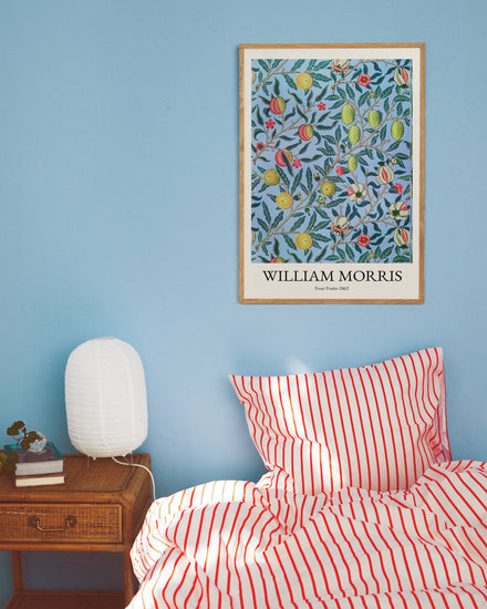 A cozy bedroom with a blue wall displays a Four Fruits by William Morris Poster. The bed has red and white striped bedding, and a wooden side table supports a white textured lamp and books, adding to the rooms charm.