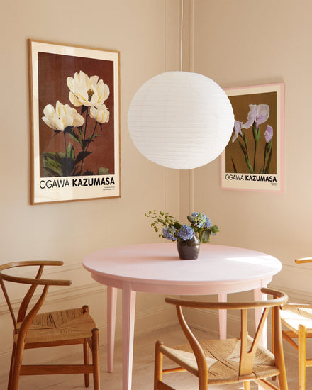 A cozy dining area with a round pink table and four wooden chairs exudes Japanese elegance, featuring a small vase of pale yellow peonies. Two Herbaceous Peony by Ogawa Kazumasa posters enhance the ambiance, illuminated by a white paper lantern above.