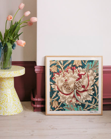 The Honeysuckle by William Morris poster, with intricate floral patterns from the Arts and Crafts era, leans against a wall. Nearby, pink tulips fill a vase on a speckled yellow and white side table. The wall and molding contrast in white and burgundy hues.