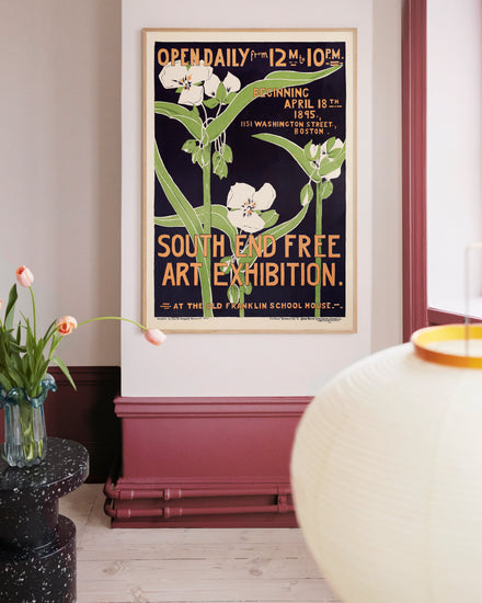 The room is adorned with a South End Free Art Exhibition 1895 Poster, set against light walls and burgundy accents. Nearby, a table holds a vase of tulips, and in the foreground, part of a round, glowing lamp is visible.