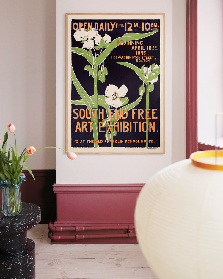 The room is adorned with a South End Free Art Exhibition 1895 Poster, set against light walls and burgundy accents. Nearby, a table holds a vase of tulips, and in the foreground, part of a round, glowing lamp is visible.