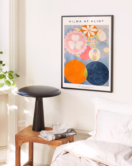 Framed artwork on a wall above a side table with a stool in a room setting.