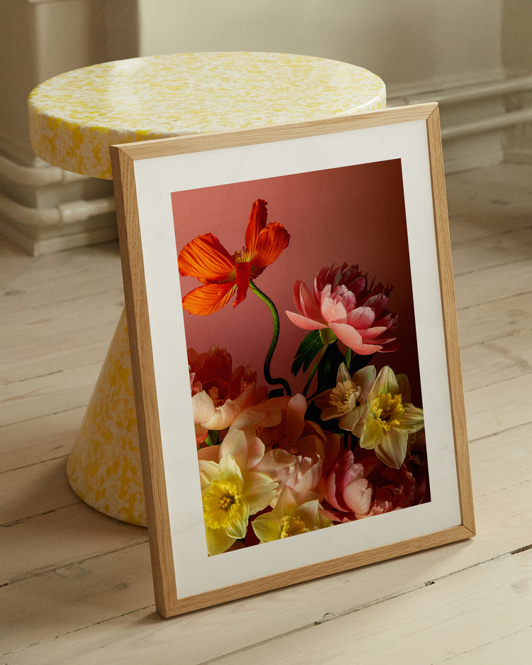 The Sprung poster showcases a vibrant still life of orange, pink peonies, and yellow flowers. It leans against a yellow-patterned stool on a light wooden floor, with soft lighting enhancing the vivid colors.