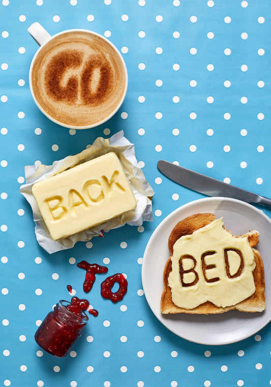 A mug of coffee spells 'GO' in froth, 'BACK' is intricately etched in butter, 'TO' is crafted from fruity jam, and 'bed' is toasted onto cheese toast. All arranged on a delightful blue tablecloth adorned with white polka dots.