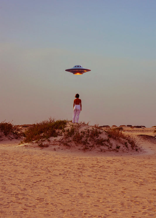Amid the deserts expanse, a person in red and white stands on a sandy hill, capturing a self-portrait with a flying saucer above. The blue sky shifts to warm hues at the horizon, evoking collage vibes in the Hey Luke, our Uber is Here! poster.