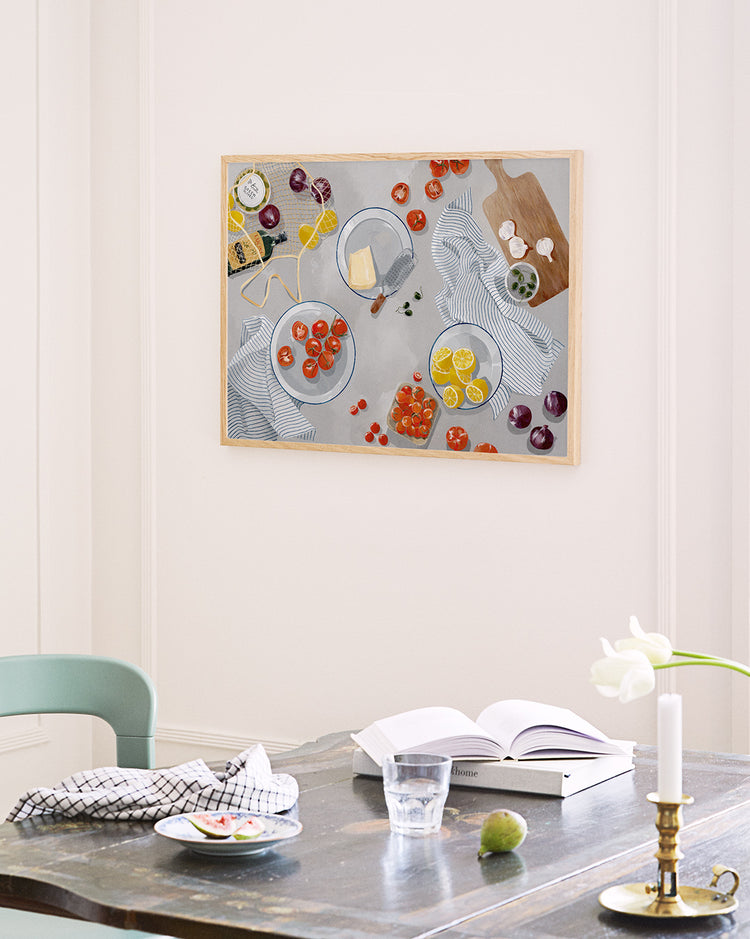 A cozy kitchen scene features a wooden table with an open book, glass of water, and candle. A green chair is partially visible. The wall displays the Like Nonna Used to Make Poster, depicting food on striped fabric. A vase with a white tulip, fresh tomatoes, and lemons enhance the inviting ambiance.