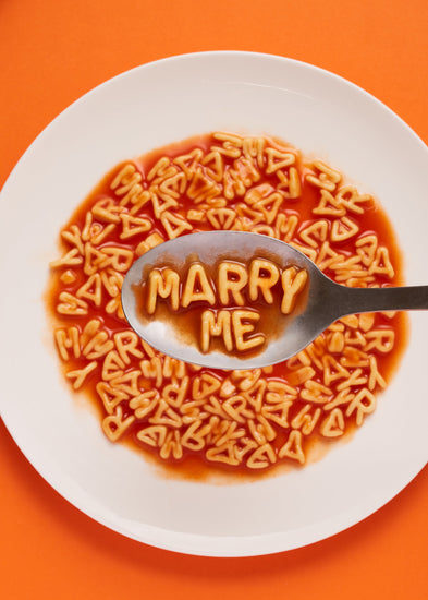 A delightful food photograph showcases alphabet spaghetti spelling out the playful phrase 'Marry Me,' creatively served on a plate with a spoon. Orange background.