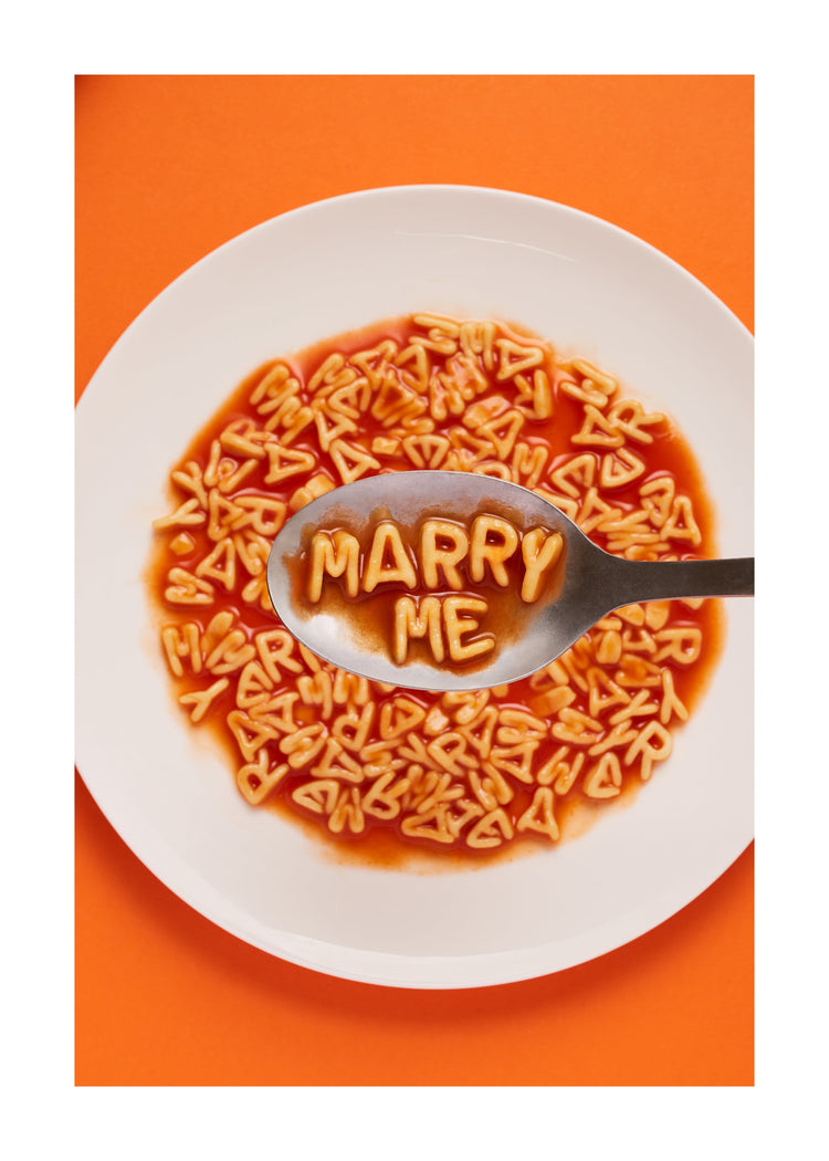 A delightful food photograph showcases alphabet spaghetti spelling out the playful phrase 'Marry Me,' creatively served on a plate with a spoon. Orange background. White border.