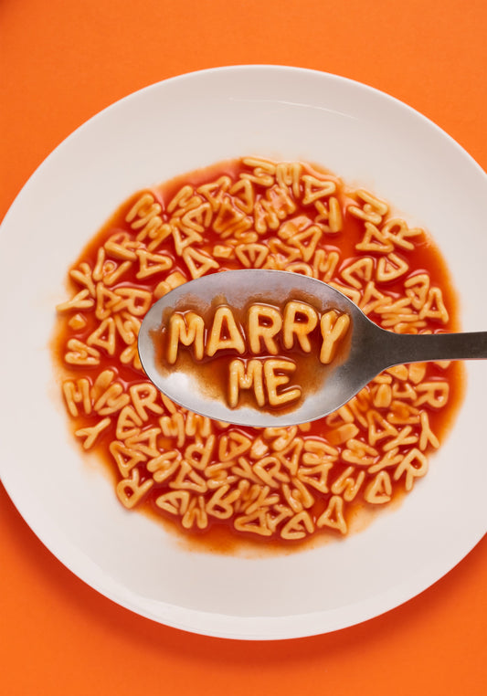 A delightful food photograph showcases alphabet spaghetti spelling out the playful phrase 'Marry Me,' creatively served on a plate with a spoon. Orange background.