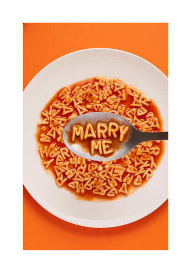 A delightful food photograph showcases alphabet spaghetti spelling out the playful phrase 'Marry Me,' creatively served on a plate with a spoon. Orange background. White border.