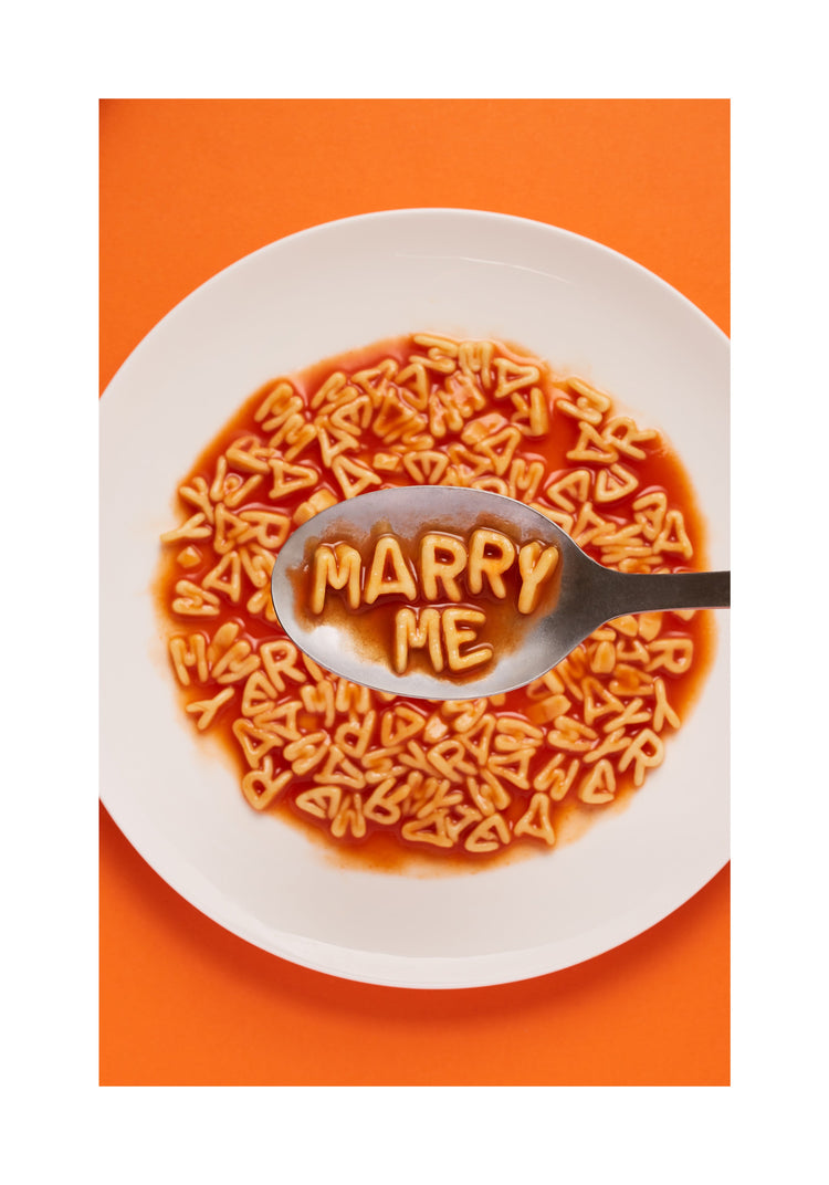 A delightful food photograph showcases alphabet spaghetti spelling out the playful phrase 'Marry Me,' creatively served on a plate with a spoon. Orange background. White border.