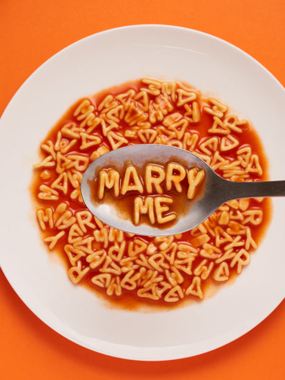 A delightful food photograph showcases alphabet spaghetti spelling out the playful phrase 'Marry Me,' creatively served on a plate with a spoon. Orange background. 