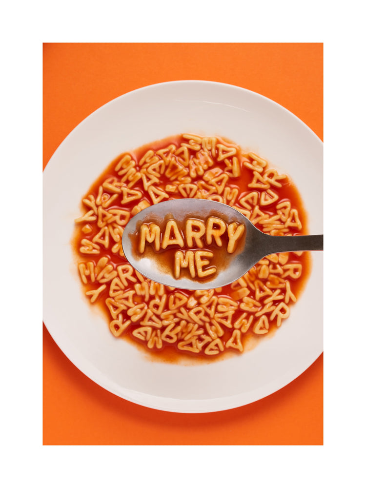 A delightful food photograph showcases alphabet spaghetti spelling out the playful phrase 'Marry Me,' creatively served on a plate with a spoon. Orange background. White border.