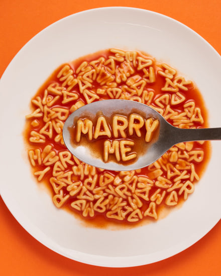 A delightful food photograph showcases alphabet spaghetti spelling out the playful phrase 'Marry Me,' creatively served on a plate with a spoon. Orange background.