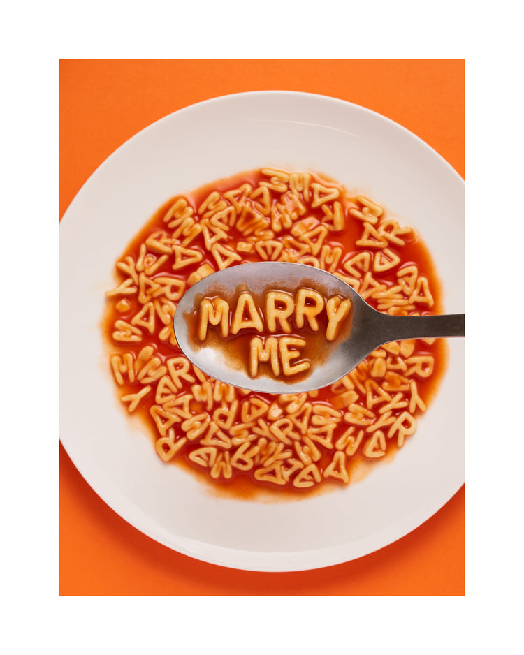 A delightful food photograph showcases alphabet spaghetti spelling out the playful phrase 'Marry Me,' creatively served on a plate with a spoon. Orange background. White border.