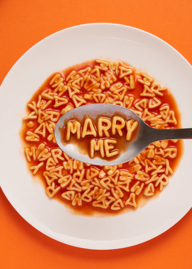 A delightful food photograph showcases alphabet spaghetti spelling out the playful phrase 'Marry Me,' creatively served on a plate with a spoon. Orange background.