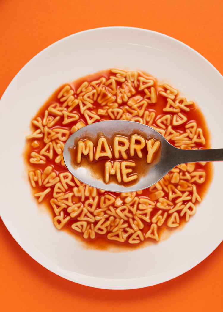 A delightful food photograph showcases alphabet spaghetti spelling out the playful phrase 'Marry Me,' creatively served on a plate with a spoon. Orange background.
