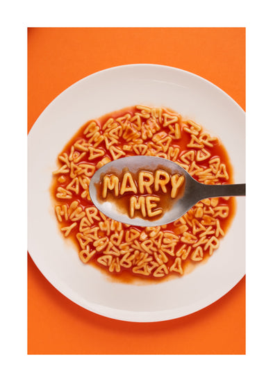 A delightful food photograph showcases alphabet spaghetti spelling out the playful phrase 'Marry Me,' creatively served on a plate with a spoon. Orange background. White border.