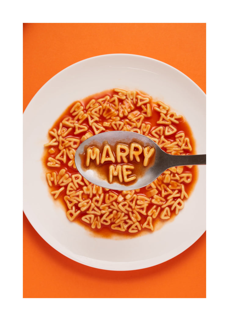 A delightful food photograph showcases alphabet spaghetti spelling out the playful phrase 'Marry Me,' creatively served on a plate with a spoon. Orange background. White border.