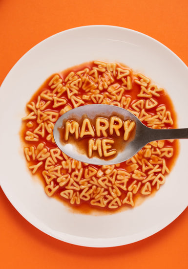 A delightful food photograph showcases alphabet spaghetti spelling out the playful phrase 'Marry Me,' creatively served on a plate with a spoon. Orange background.