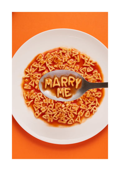 A delightful food photograph showcases alphabet spaghetti spelling out the playful phrase 'Marry Me,' creatively served on a plate with a spoon. Orange background. White border.