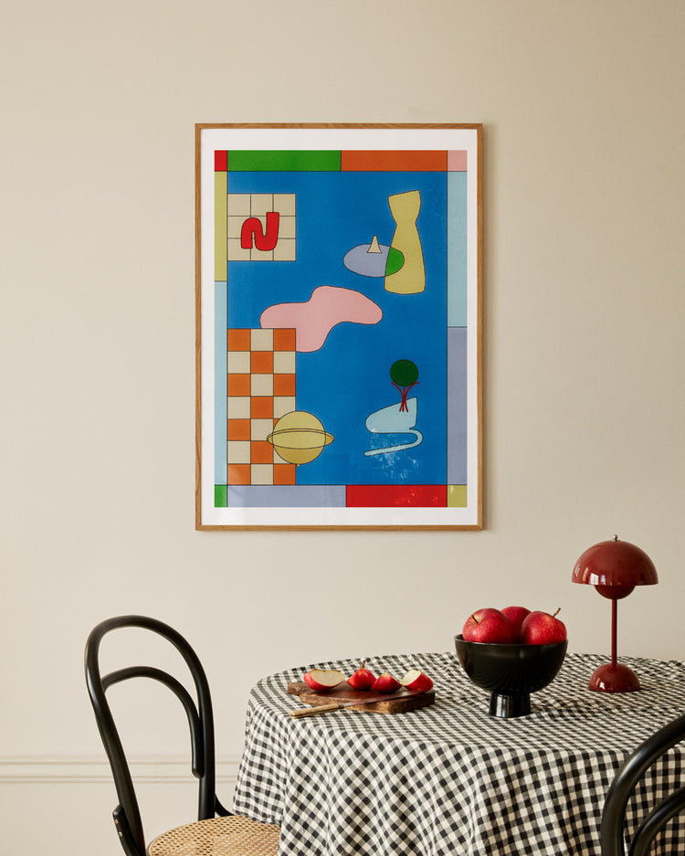 A Space Lamp Poster featuring colorful shapes decorates the neutral wall. Below it, a round table with a black and white checkered cloth holds a black bowl of apples and a red lamp, complemented by two black chairs with woven seats around the table.