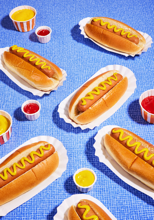 Still life photograph of perfectly grilled hot dogs adorned with mustard and served on a vibrant blue table cloth.