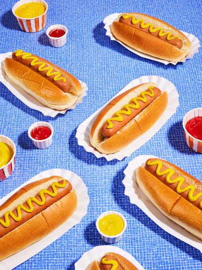 Still life photograph of perfectly grilled hot dogs adorned with mustard and served on a vibrant blue table cloth.