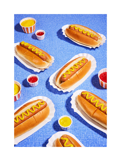Still life photograph of perfectly grilled hot dogs adorned with mustard and served on a vibrant blue table cloth. White border.