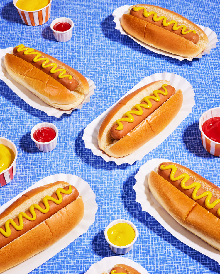 Still life photograph of perfectly grilled hot dogs adorned with mustard and served on a vibrant blue table cloth.