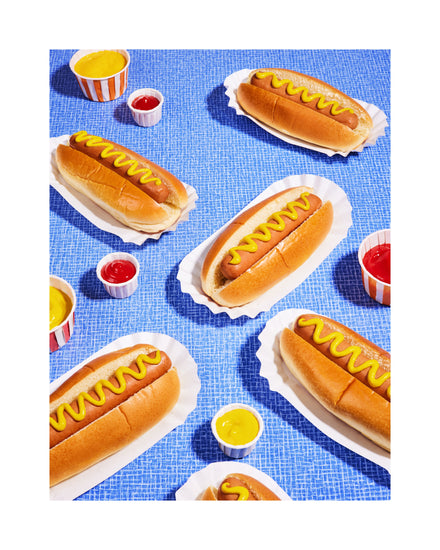Still life photograph of perfectly grilled hot dogs adorned with mustard and served on a vibrant blue table cloth. White border.