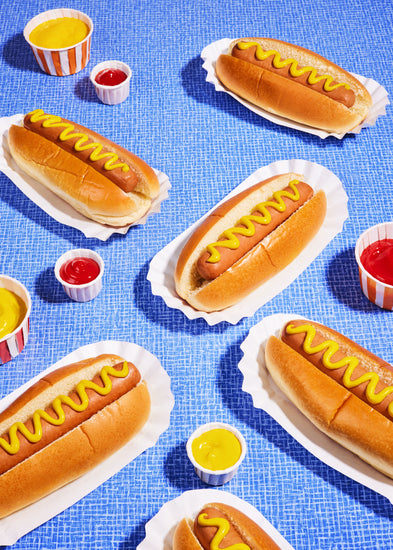 Still life photograph of perfectly grilled hot dogs adorned with mustard and served on a vibrant blue table cloth.