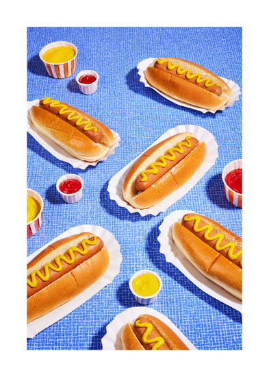 Still life photograph of perfectly grilled hot dogs adorned with mustard and served on a vibrant blue table cloth. White border.