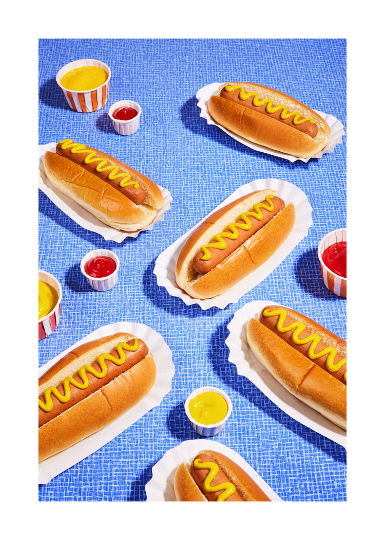 Still life photograph of perfectly grilled hot dogs adorned with mustard and served on a vibrant blue table cloth. White border.