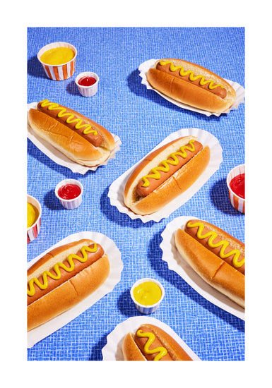 Still life photograph of perfectly grilled hot dogs adorned with mustard and served on a vibrant blue table cloth. White border.