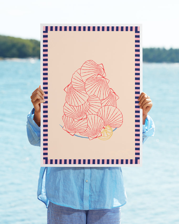 In a blue shirt and striped pants, someone holds the Saint-Jacques du Marché Poster, featuring abstract red line art of seashells in collage style with a small yellow lemon. A body of water and trees appear in the background, while the poster obscures their face.
