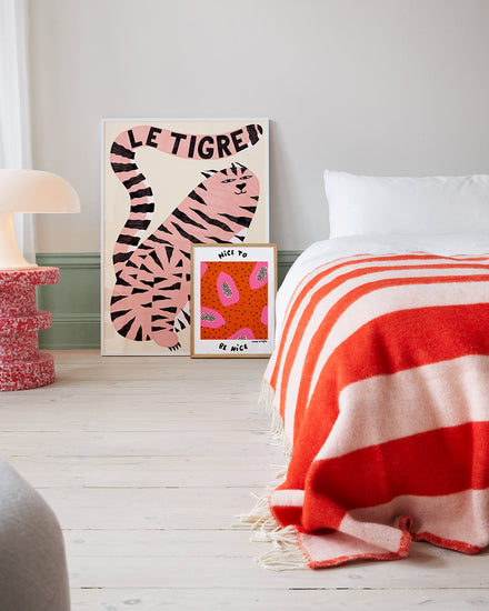 A minimalist bedroom exudes wild sophistication with a red and white striped bedspread. Two abstract art pieces lean against the wall: a large one showcasing Le Tigre White Poster with a pink tiger illustration and a smaller one with bold shapes. A modern lamp sits on a textured red side table.